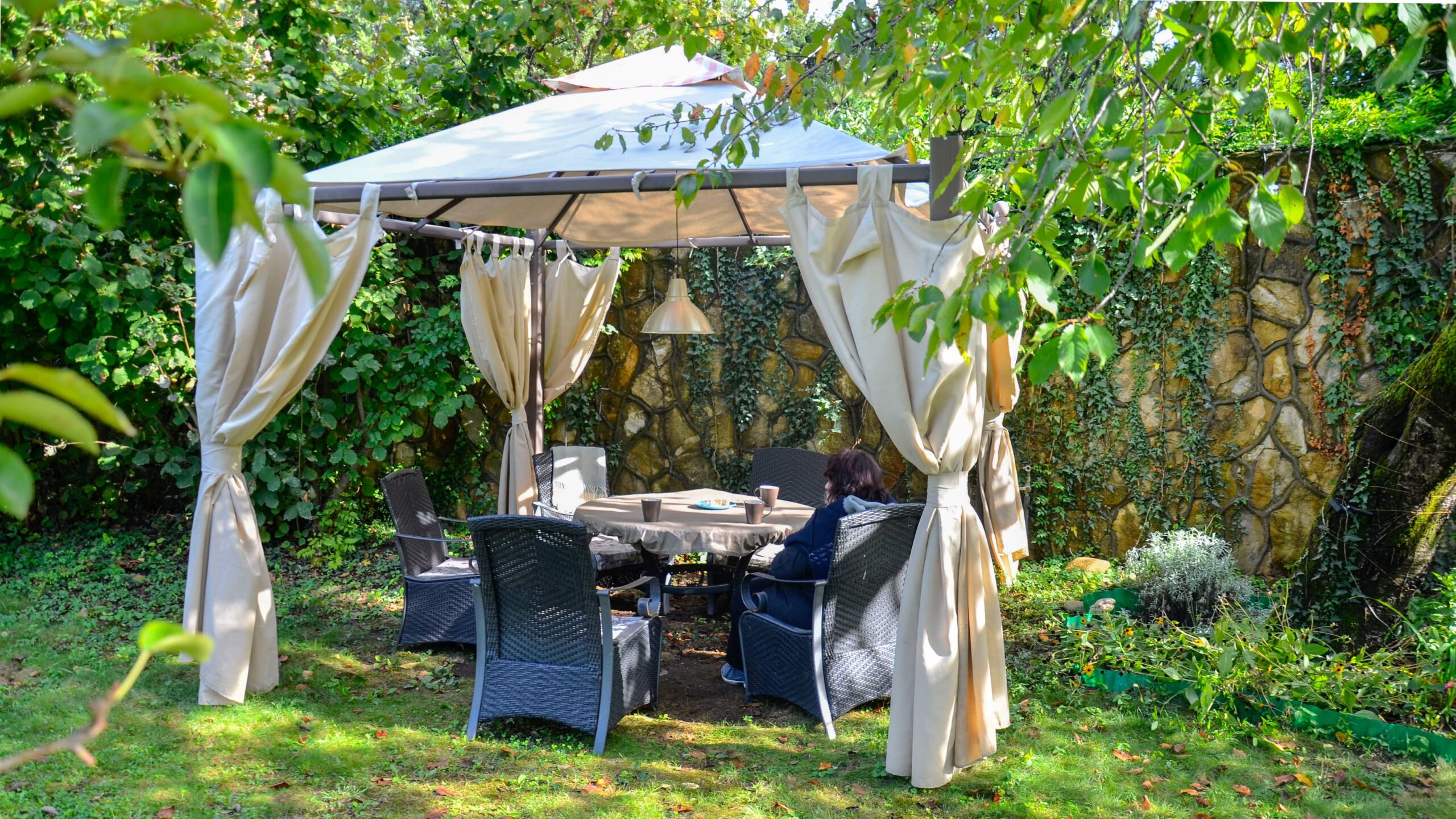 Gartenzelt mit Stoffvorhängen, Rattan Stühlen und hängender Lampe in grüner Gartenecke